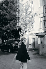 A woman in a stylish black coat stands in a street scene with modern architecture