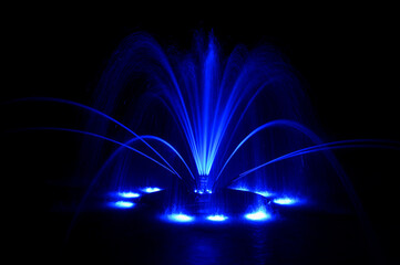 Floating fountain in neon blue, large detailed colorful horizontal closeup at night, lit up illuminated color changing bright city park lights, isolated colored water jets on dark black background