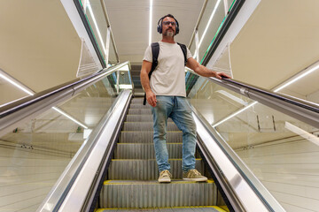 Middle-Aged Man in Casual Clothing with Backpack Descending on Escalator at Train Station to Reach...
