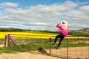 Woman farmer sits on a rusty gate fence overlooking canola farm fields