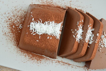Dark chocolate milk pudding cake with cocoa powder and coconut flakes topping served on a plate. Selective focus