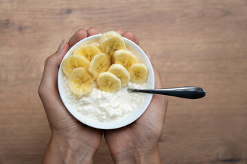 Woman holding a bowl of cottage cheese and banana for a healthy breakfast or snack on a wooden table with copy space