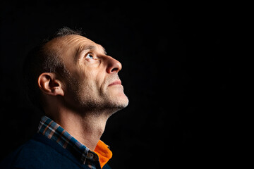 Fototapeta premium Older man with a neat beard and short gray hair looks upwards in deep contemplation against a dark backdrop, exuding wisdom and thoughtfulness