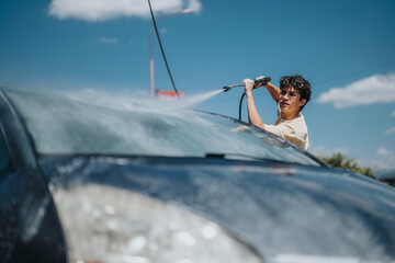A young man uses a high-pressure hose to wash his car on a sunny day. The vibrant scene captures...