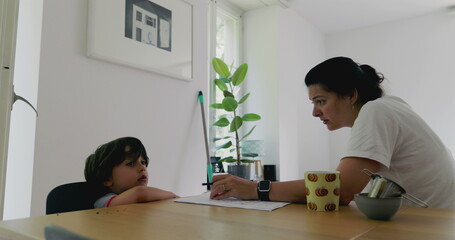 Mother struggling to homeschool her son at a desk, focusing on educational activities in a bright and airy living space