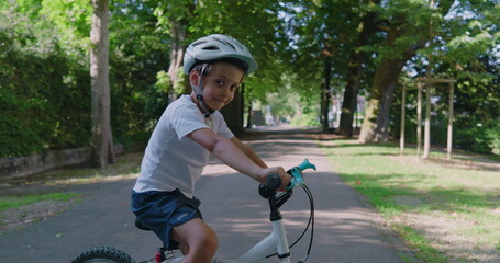 Smiling boy on a bicycle in a park, wearing a helmet for safety, paused on a path lined with trees,...