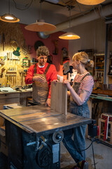 Carpenter colleagues, man and woman, discuss work details in workshop. Male mentor explains...