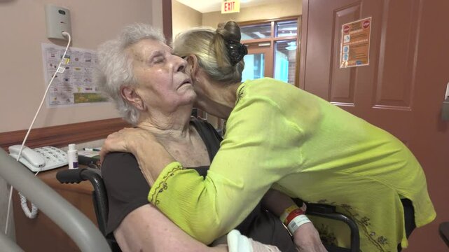An elderly woman with white hair sits in a wheelchair and receives a hug and kiss on the cheek from a woman wearing a yellow shirt. They are in an indoor setting, likely a care facility.