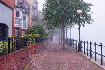 Obraz premium Foggy Morning Along a Quiet Riverside Path Lined with Brick Apartments, Trees, and Classic Streetlamps, Creating a Serene Urban Scene Disappearing into the Mist
