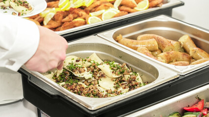 A person in a white uniform scoops a heaping serving of pierogi with beacon from a stainless steel chafing dish