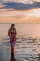 A beautiful young woman in a pink dress is standing on the seashore at sunset.