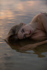 A beautiful woman in lingerie is lying in the water on the seashore at sunset. Portrait