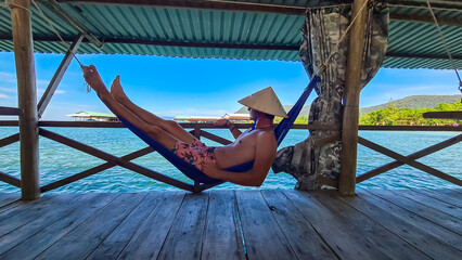 Man relaxing in hammock on wooden platform overlooking a serene coastal scene in Rach Vem, Phu Quoc island, Vietnam, South East Asia. He is wearing straw hat and enjoying view of calm turquoise sea