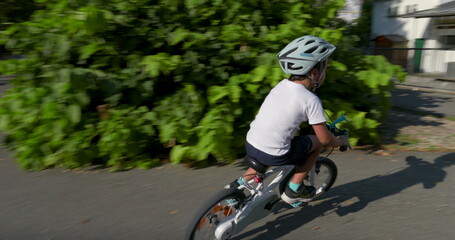 Obraz premium Young boy crossing a crosswalk on his bicycle, wearing a helmet for safety, in an urban setting with trees and street signs, enjoying outdoor activity on a sunny day