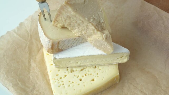 Medium hard cheese heads brie, parmesan on craft paper. Pile pieces of different kinds handmade cheese stacked in vertical row on wooden table with cheese fork on top - healthy organic eating concept