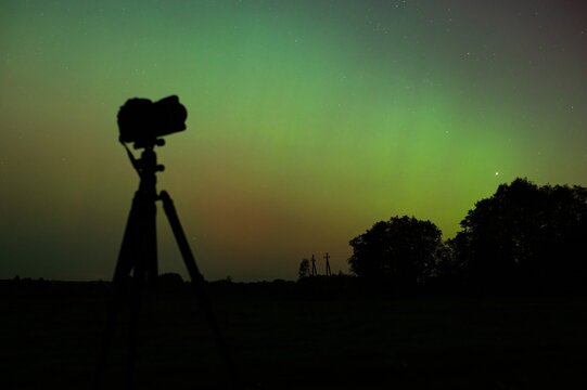 World Photography Day. Photographer with his camera and tripod taking pictures of Northern Lights. Aurora Borealis over fields landscape. Lithuania 10 10 2024