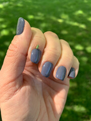 A close-up of a woman's bent hand with purple nails, holding a small insect on one finger. The background is washed out greenery, making the tiny insect the main focus of the image