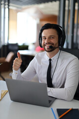 A man wearing a headset and a black tie is sitting at a desk with a laptop. He is smiling and...