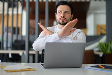 A man is sitting at a desk with a laptop in front of him. He is making a hand gesture that looks...