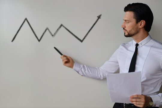 A man is standing in front of a whiteboard with a graph on it. He is holding a marker and pointing to the graph. The graph shows a line that is going up, which could represent an increase in something