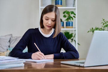 Young serious woman studying working writing in notebook, sitting at home at desk