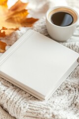 white blank book on top of cosy wool blanket, dry maple leaves, cup of coffee with latte art, top view, warm autumn colors, sun and shadows