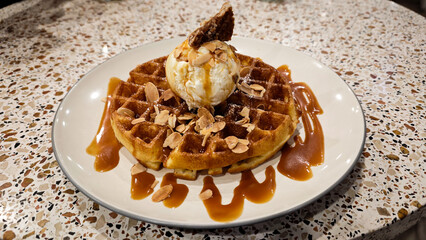 Closeup of a golden brown waffle topped with vanilla ice cream, almond flakes, and caramel drizzle on a terrazzo-style table at a modern cafe