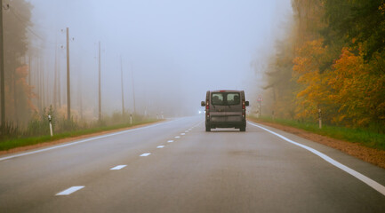 A Van Driving Along a Foggy Road Surrounded by Autumn Foliage in a Tranquil Rural Setting During the Early Morning Hours