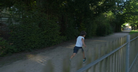 Young boy running down a tree-lined path, wearing a white shirt and shorts, surrounded by greenery, enjoying an energetic moment outdoors on a sunny day