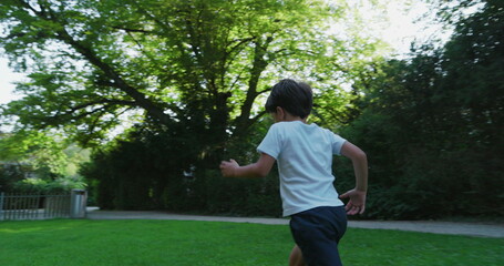 Young boy running through a grassy park, viewed from behind, wearing a white shirt and shorts, surrounded by tall trees and greenery, enjoying an active day outdoors