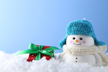 Cute decorative snowman and gift box on artificial snow against light blue background, closeup. Space for text