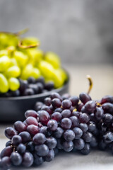 Bunches of black and green grapes lying on a light surface. In the background, a bowl with green and black grapes is visible.