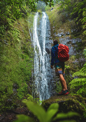 Male tourist carrying a rain-covered backpack stands in front of waterfall. Poco dos Pulgas waterfalls, Madeira Island, Portugal, Europe.