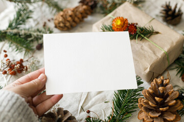 Christmas card mock up. Hand holding empty greeting card on rustic table with fir branches and pine...