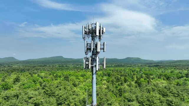 4G/5G cell tower with telecom antennas for several companies high above a lush green forest in New England aerial drone video push-in