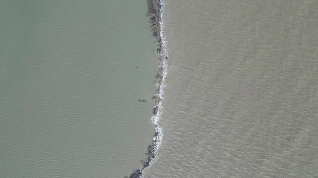 Aerial view of small sea waves crashing against a barrier, with water on both sides. The scene captures the continuous movement of the waves as they meet the barrier, creating a dynamic and serene coa