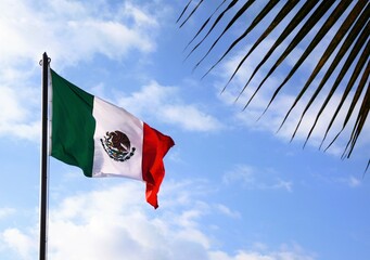 The Mexican flag flies up a flagpole on a beautiful summer day with a background of blue sky with...