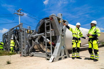 Fototapeta premium Two engineers in safety gear inspecting a wind turbine blade section on a construction site. They examine the metal framework with precision. Renewable energy project under a clear blue sky.