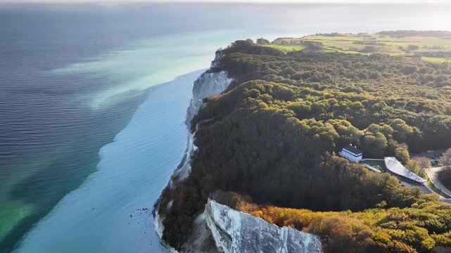 View of white cliffs, dense forest and building in classic Scandinavian architectural style perched near the edge against the backdrop of a calm blue sea. Landscape of chalk cliffs Mons Klint.