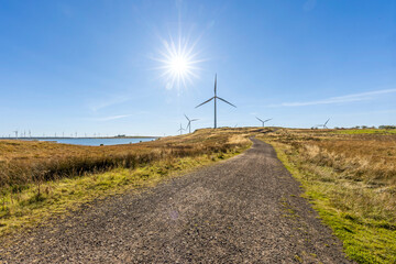 Gravel dirt track road leading to freshwater lake and wind farm turbines on grassy moor land sunburst in sky