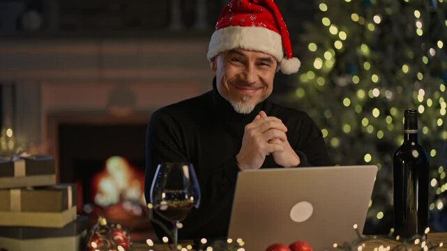 Portrait of happy middle aged man in Santa hat at Christmas, smiling, sitting at desk in front of fireplace at home with laptop computer and glass of red wine. Holiday greeting.