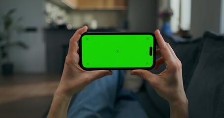 A woman holds a mobile phone with green screen in her hand. She is lying on the couch at home, close-up of her hands, unrecognizable person