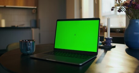 An open laptop with a green screen is on the table in the kitchen at home. A cup of hot drink nearby