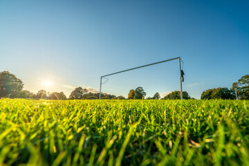 Low angle view of football or soccer goal post on a green grass pitch in a park at sunset time. Sport theme background