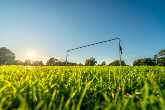 Low angle view of football or soccer goal post on a green grass pitch in a park at sunset time. Sport theme background