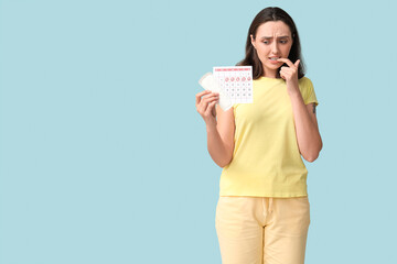 Beautiful displeased woman holding pad and calendar with marked days of menstruation on blue background