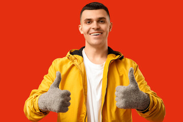 Young man in stylish jacket and gloves showing thumb-up on red background