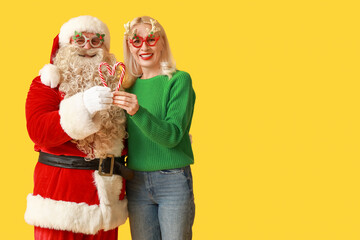 Santa Claus and beautiful mature woman in Christmas glasses with candy canes on yellow background