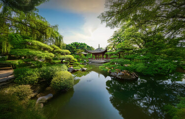Fototapeta premium A traditional Chinese-style building with a red roof sits on the edge of a tranquil pond surrounded by lush greenery.