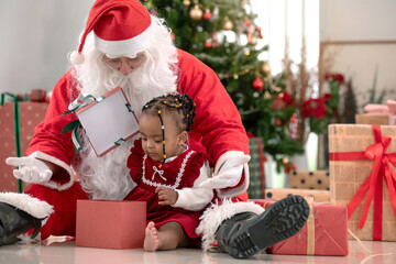 Little African girl in a red dress sits on the floor with Santa Claus near Christmas tree at home, paying attention to the gift box in his hand, celebrate Christmas